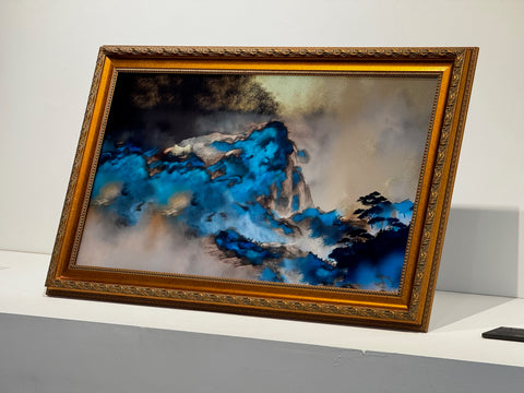 這張照片展示一幅新東方山水畫作,畫面採用濃烈的青金石藍與金黃暈染,營造出層次分明、煙霧繚繞的山嵐意境。畫作以仿古金色雕花木框裝裱,整體色澤厚重高雅,細緻的紋路襯托出藝術品的珍貴。這幅畫特別在友達AUO FineARTs 32吋擬真藝屏上展出,電子螢幕還原畫作的光澤與細節,讓色彩與筆觸都能逼真呈現於現場,完美結合傳統與科技,適合藝術愛好者收藏與欣賞。